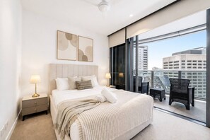Quarto de hotel com uma cama grande, duas mesas de cabeceira com luminárias, uma varanda com cadeiras e vista para a cidade.