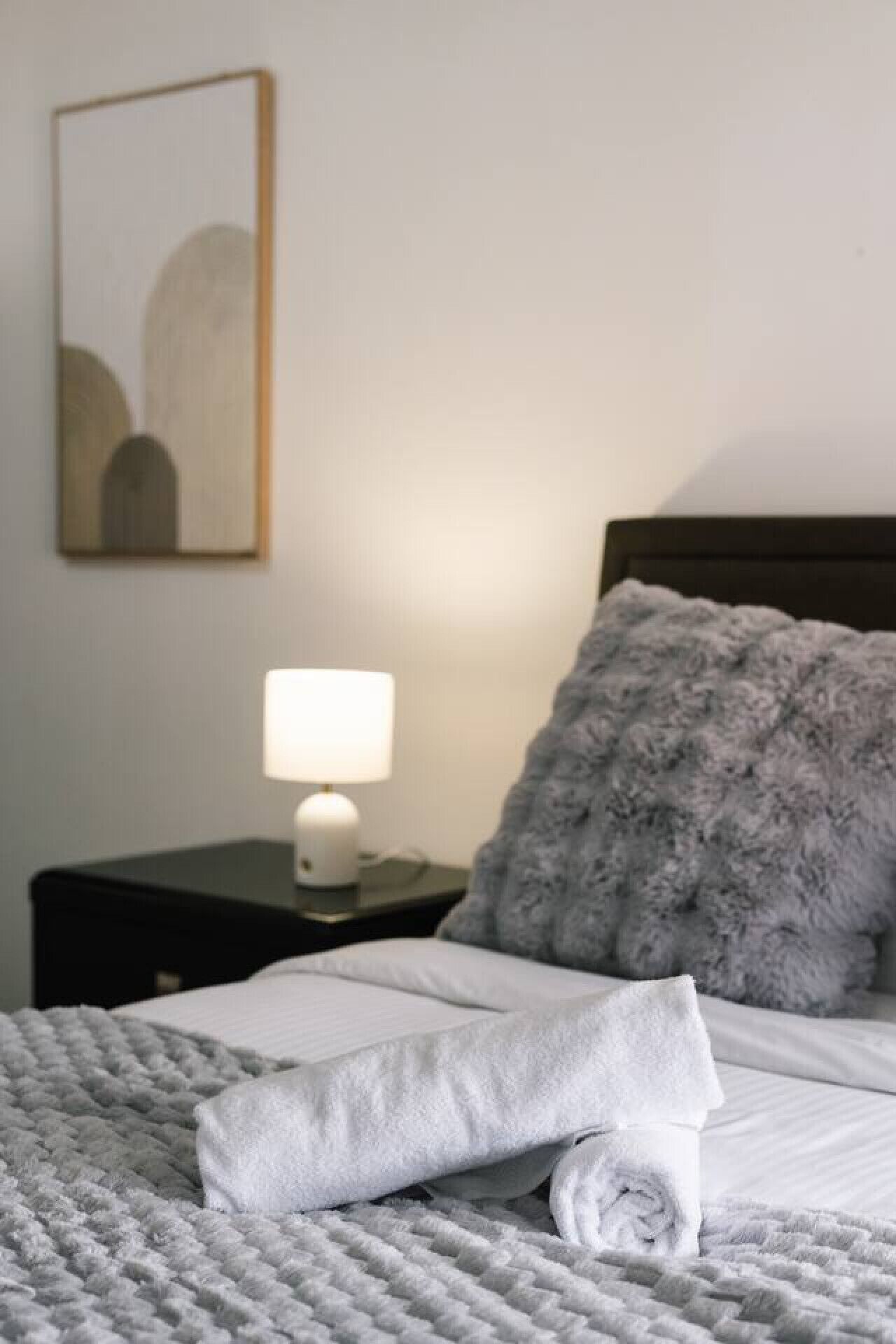 Un lit bien fait, avec une serviette roulée, un oreiller moelleux, une lampe de chevet et un tableau encadré accroché au mur.