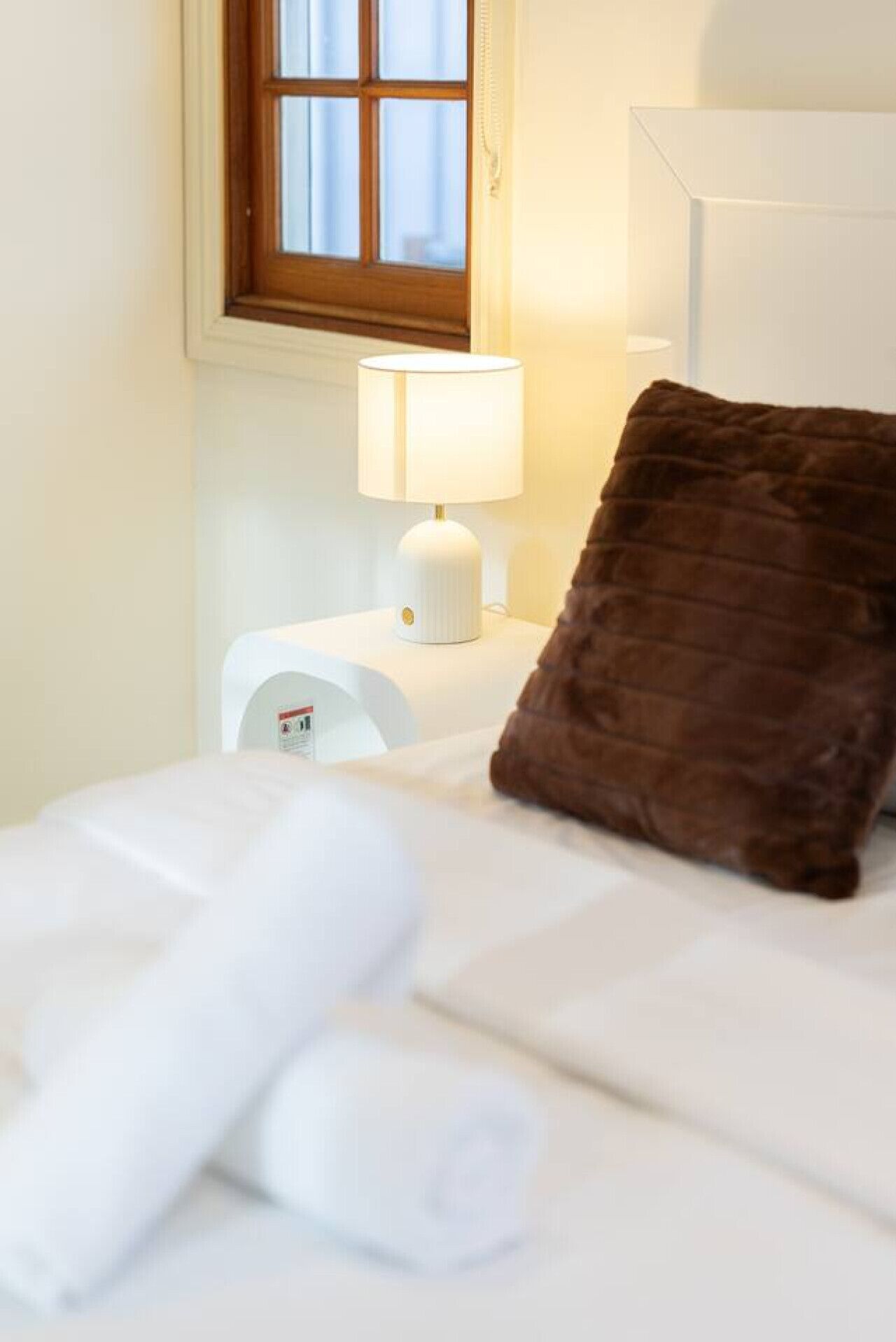 Een net opgemaakt bed met witte lakens, een bruin kussen en een nachtkastje met een lamp.