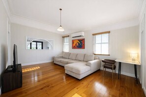 Una sala de estar con sofá, televisión, escritorio con lámpara y un cuadro en la pared.