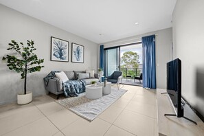 A modern living room with a grey sofa, a coffee table, a TV, a balcony with blue curtains, and a potted plant. - 2BR Apartment near Sunnybank Plaza & Market Square (MacGregor)