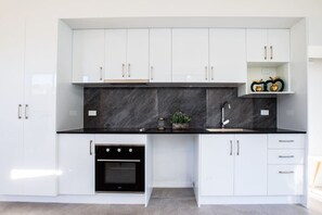 A modern kitchen with white cabinets, a black countertop, a built-in oven, and a small plant on the counter. - Budget Private Suite w/ Kitchen, Ensuite & A/C (Oxley)