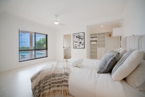 Une chambre à coucher comprenant un lit, une fenêtre avec des stores, un ventilateur de plafond, un tableau encadré et un dressing.