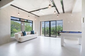 A modern living room with a white sofa, a pool table, and large windows offering a view of greenery. - Luxury 6BR Hinterland Retreat w/ Pool &Scenic View (Bonogin)