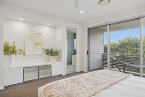 A bedroom with a bed, a bench, two potted plants, a framed artwork, and a sliding glass door leading to an outdoor area.