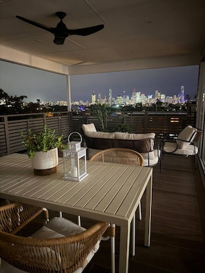 A balcony with a table, chairs, a ceiling fan, and a cityscape view at night.
