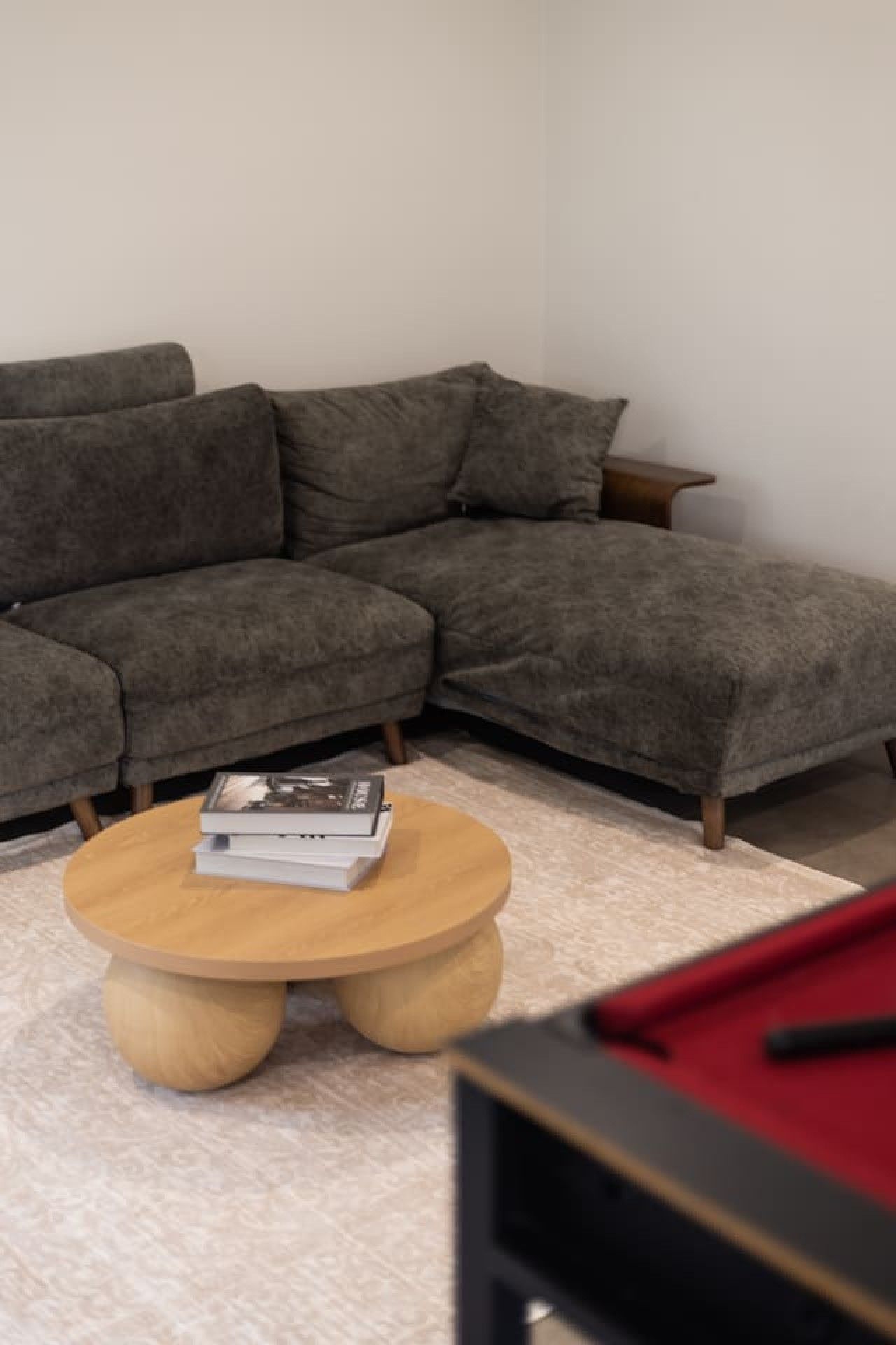 A living room with a grey sectional sofa, a round wooden coffee table with books, and a red pool table.