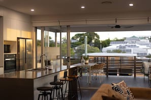 A modern kitchen with a large island, bar stools, and a dining area with a view of buildings and greenery. - Scenic City Views + Pool | Elegant 5BR Residence (Hawthorne)