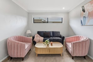 A living room with a dark grey sofa, two pink armchairs, a wooden coffee table, and a window with blinds.