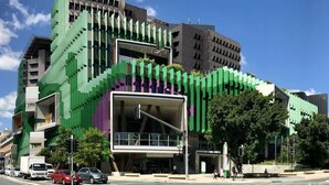 Ein modernes Gebäude mit einer auffälligen, grün-violetten Fassade, mehreren Balkonen und einem Fußgängerbereich mit Bäumen.
