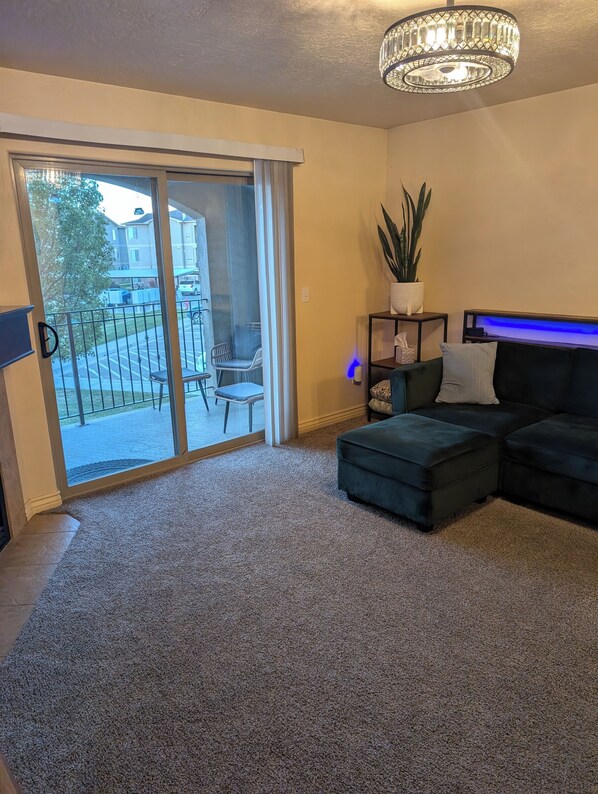 A living room with a sliding glass door to a balcony, a dark sofa, a small table with a plant, and a blue-lit shelf.