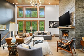 A modern living room with a fireplace, large windows, and a flat-screen TV. - Moonlight Creek Retreat ~ Private Creek, Hot Tub, Fire Pit & More (Ellijay)