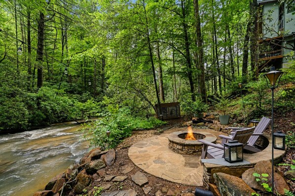A stone fire pit with seating and lanterns in a forested area with a stream. - Moonlight Creek Retreat ~ Private Creek, Hot Tub, Fire Pit & More (Ellijay)