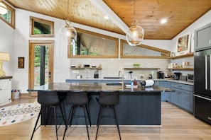 A modern kitchen with a large island, bar stools, a refrigerator, and a view of greenery outside. - Moonlight Creek Retreat ~ Private Creek, Hot Tub, Fire Pit & More (Ellijay)