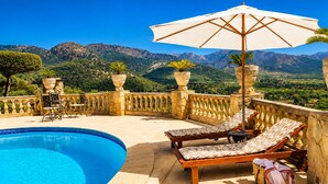 Zona de piscina amb gandules, una taula i un parasol gran.