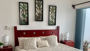 Ein Schlafzimmer mit rotem Kopfteil, einem großen Bett mit weißer Bettwäsche, drei gerahmten Pflanzenbildern an der Wand, zwei Nachttischen mit Lampen und einem Fenster mit Blick.