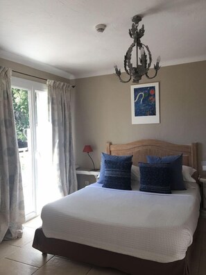 A bedroom with a bed, curtains, a chandelier, a nightstand, a lamp, and a painting on the wall. - Hotel les Messugues By Alain Llorca (Saint-Paul-de-Vence)