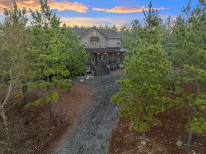 Eine zweistöckige Holzhütte mit Veranda und einem Schotterweg, umgeben von Kiefern.