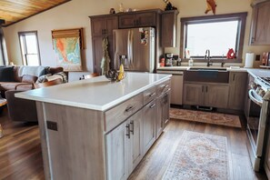 A modern kitchen with a central island, wooden cabinets, a stainless steel refrigerator, and a large window.