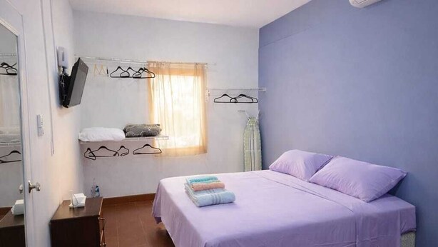A bedroom with a bed, a small bedside table, a mirror, a wall-mounted TV, and a window with curtains.
