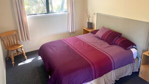Une chambre à coucher comprenant un lit, une chaise, une table de chevet, une fenêtre avec des rideaux et une vue sur les arbres.