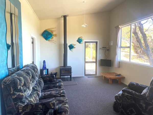 A living room with a sofa, a wooden table, a fireplace, and a large window with a view of trees. - Kupe's Secret (The Bush Bach) (Palliser Bay)