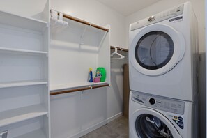 A laundry room with a stacked washer and dryer, a drying rack, and a shelf with cleaning supplies. - Blue Desert Oasis - 4 Bed w/ Bonus Casita and Pool and Firepit (Goodyear)