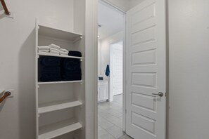Un placard blanc avec des étagères et une tringle pour suspendre les vêtements, une porte blanche, et on aperçoit un coin d’une salle de bain avec un porte-serviettes.