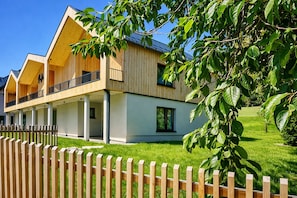 Ein modernes, zweistöckiges Holzhaus mit Balkon, umzäunter Garten und Baum im Vordergrund.