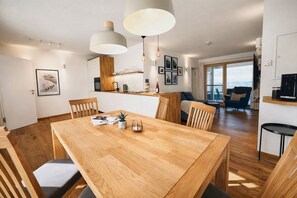 Área de refeições moderna com mesa de madeira, cadeiras, luminárias pendentes e vista para o mar através de uma grande janela.