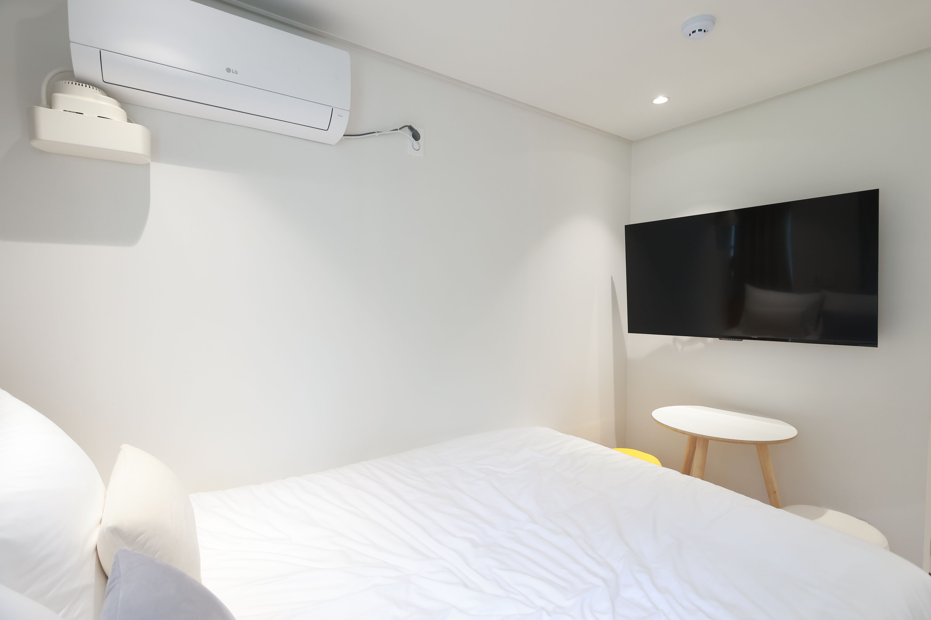 A modern hotel room with a bed, a flat-screen TV mounted on the wall, a small round table, and a wall-mounted air conditioning unit.