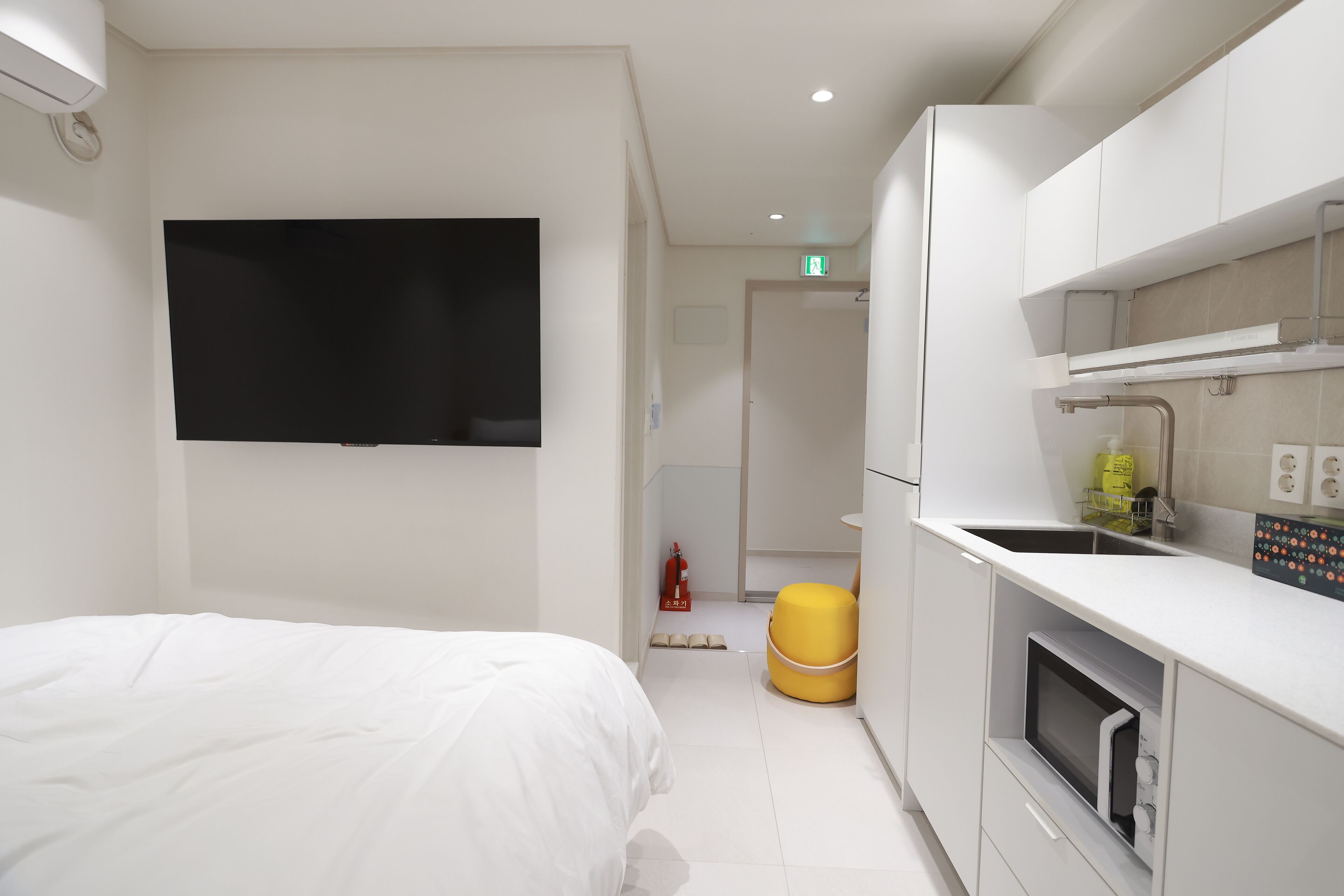 A modern hotel room with a bed, a TV, a kitchen area with a microwave, and a yellow ottoman.