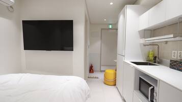 A modern hotel room with a bed, a TV, a kitchen area with a microwave, and a yellow ottoman.