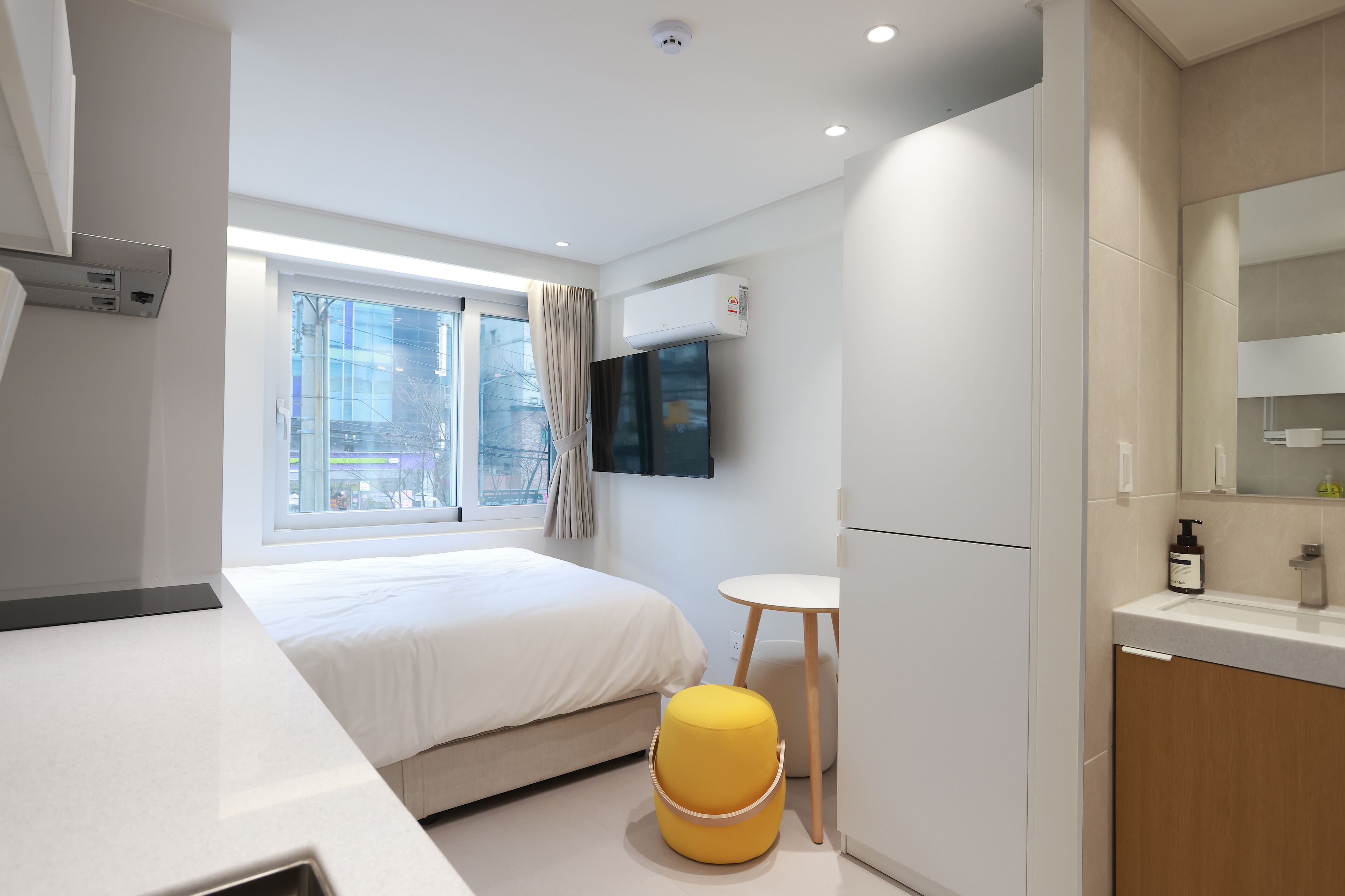 A modern hotel room with a bed, a small round table, a yellow ottoman, a TV mounted on the wall, and a window with curtains.