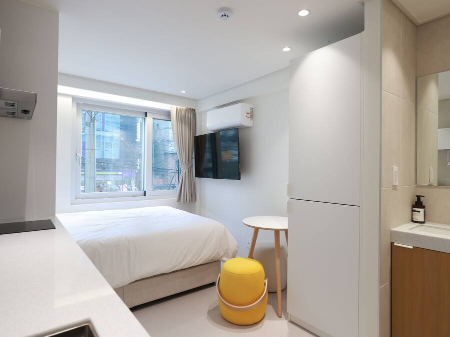 A modern hotel room with a bed, a small round table, a yellow ottoman, a TV mounted on the wall, and a window with curtains.