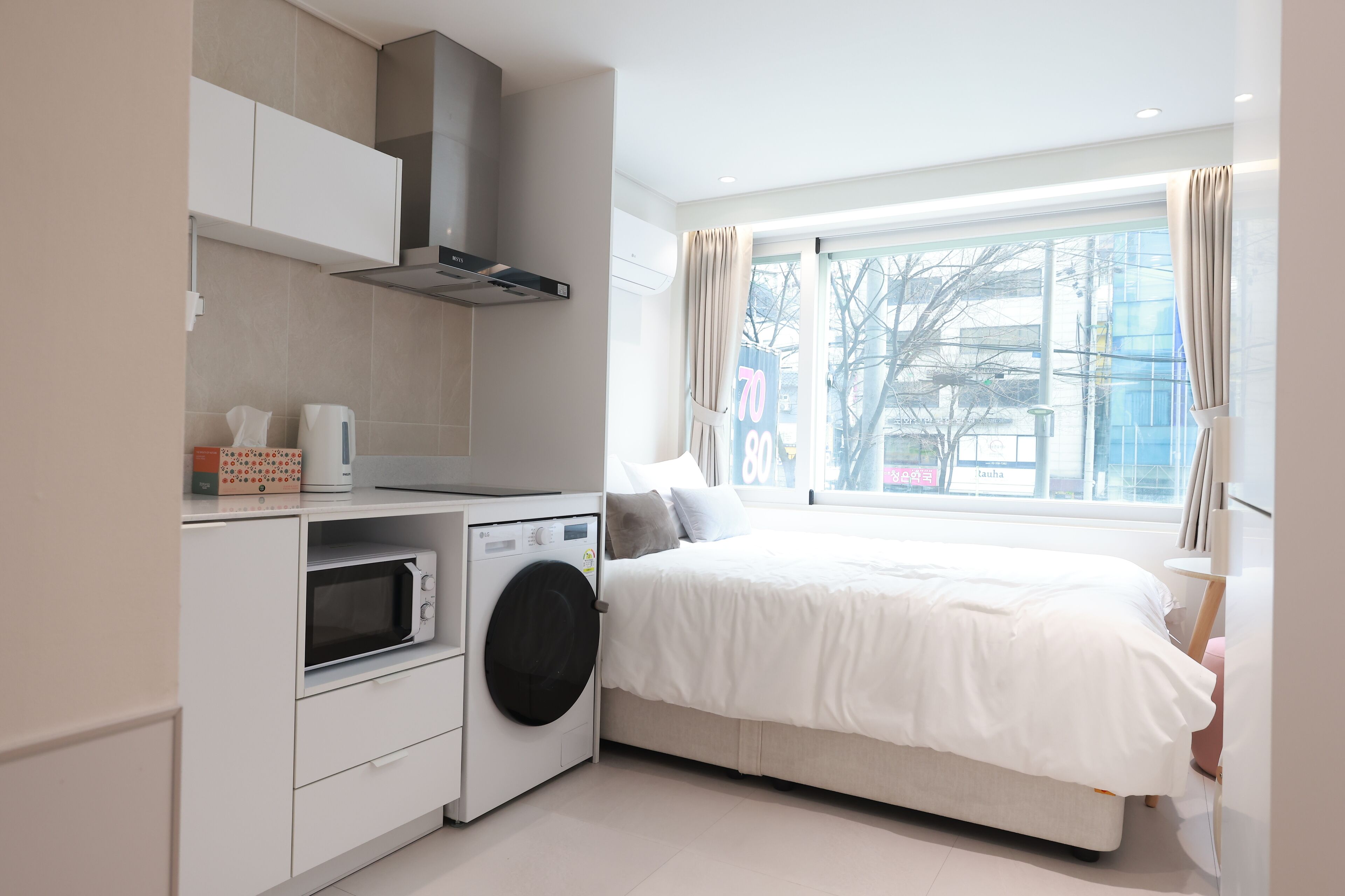 Ein kleines Hotelzimmer mit Bett, Waschmaschine, Mikrowelle und einem fenster mit Vorhängen.