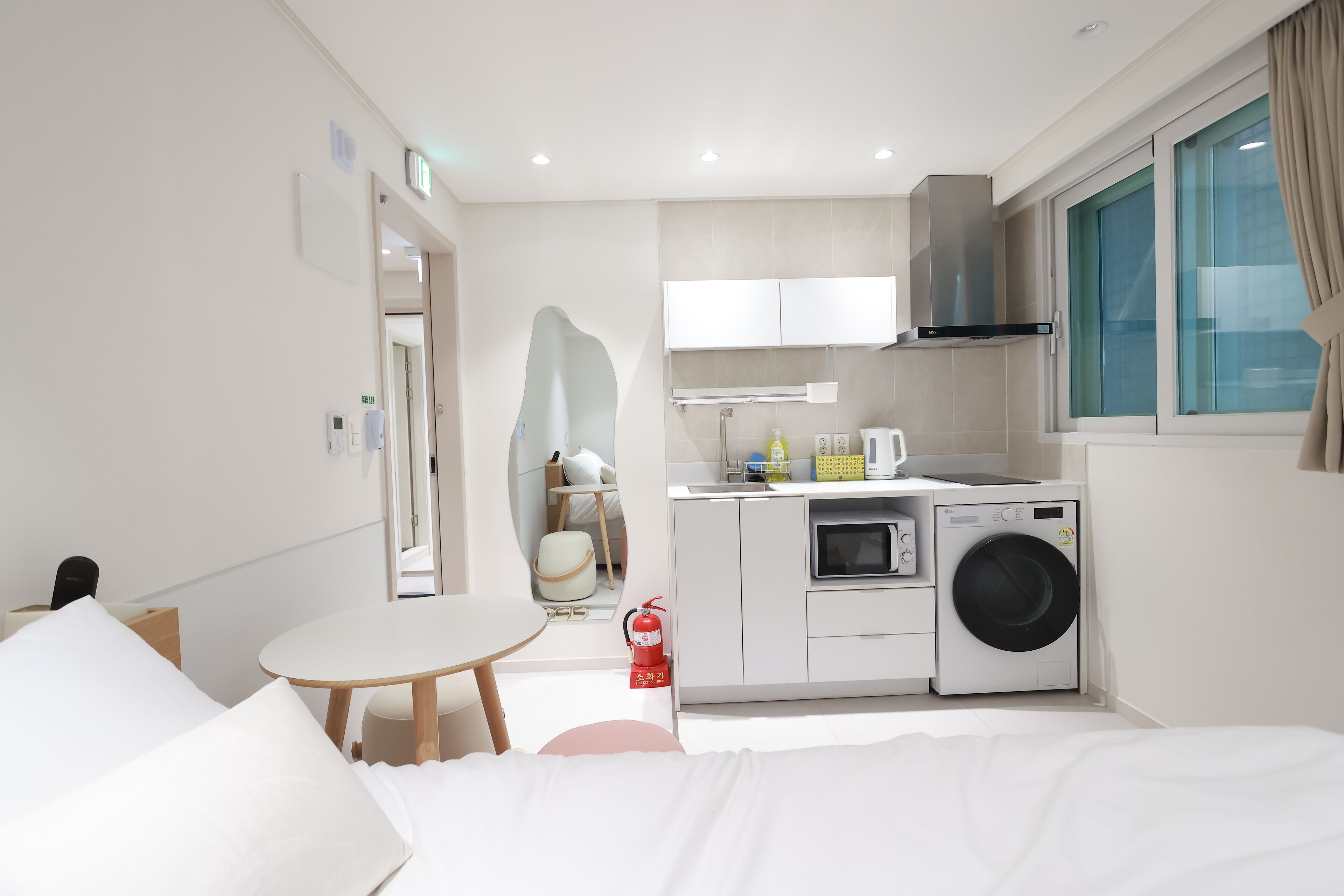 A modern hotel room with a bed, a small table, a chair, a washing machine, a microwave, and a kitchenette.