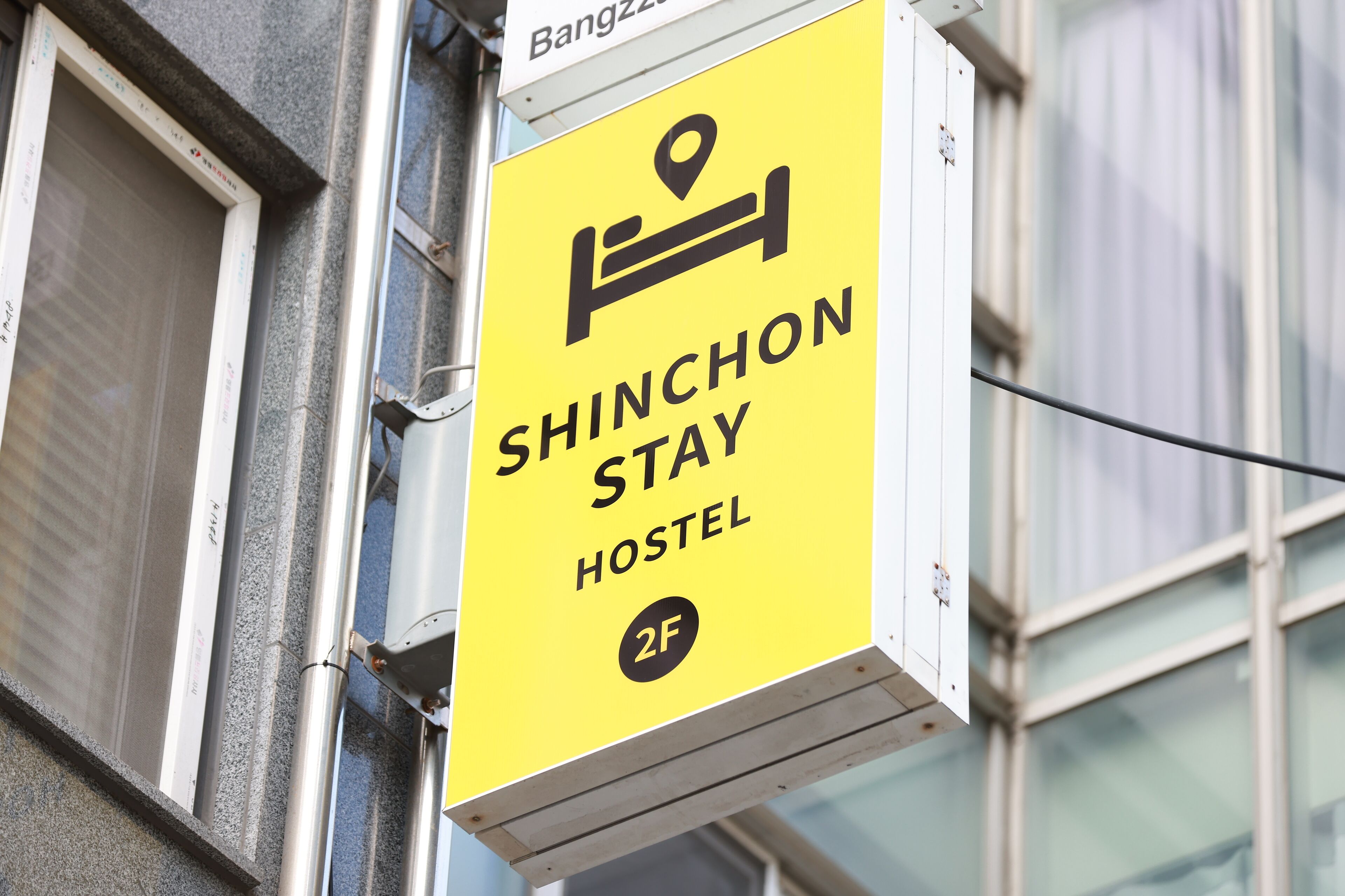Une enseigne jaune indiquant un auberge de jeunesse, avec un symbole de lit et le texte « SHINCHON STAY HOSTEL 2F ».