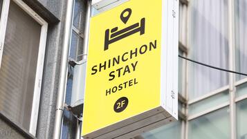 Une enseigne jaune indiquant un auberge de jeunesse, avec un symbole de lit et le texte « SHINCHON STAY HOSTEL 2F ».