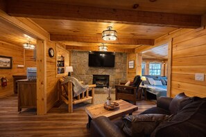A cozy log cabin interior with a stone fireplace, wooden furniture, and a television mounted on the wall. - Whiskey N Wine Ridge: Fire Pit, Hot Tub, Ample Parking, pets, near Pigeon Forge (Sevierville)
