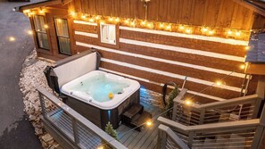 A hot tub on a wooden deck with string lights, a wooden cabin, and a stone wall. - Whiskey N Wine Ridge: Fire Pit, Hot Tub, Ample Parking, pets, near Pigeon Forge (Sevierville)