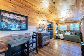 Acogedor interior de cabaña con techo de madera, zona de comedor con mesa y sillas, sofá y televisor.