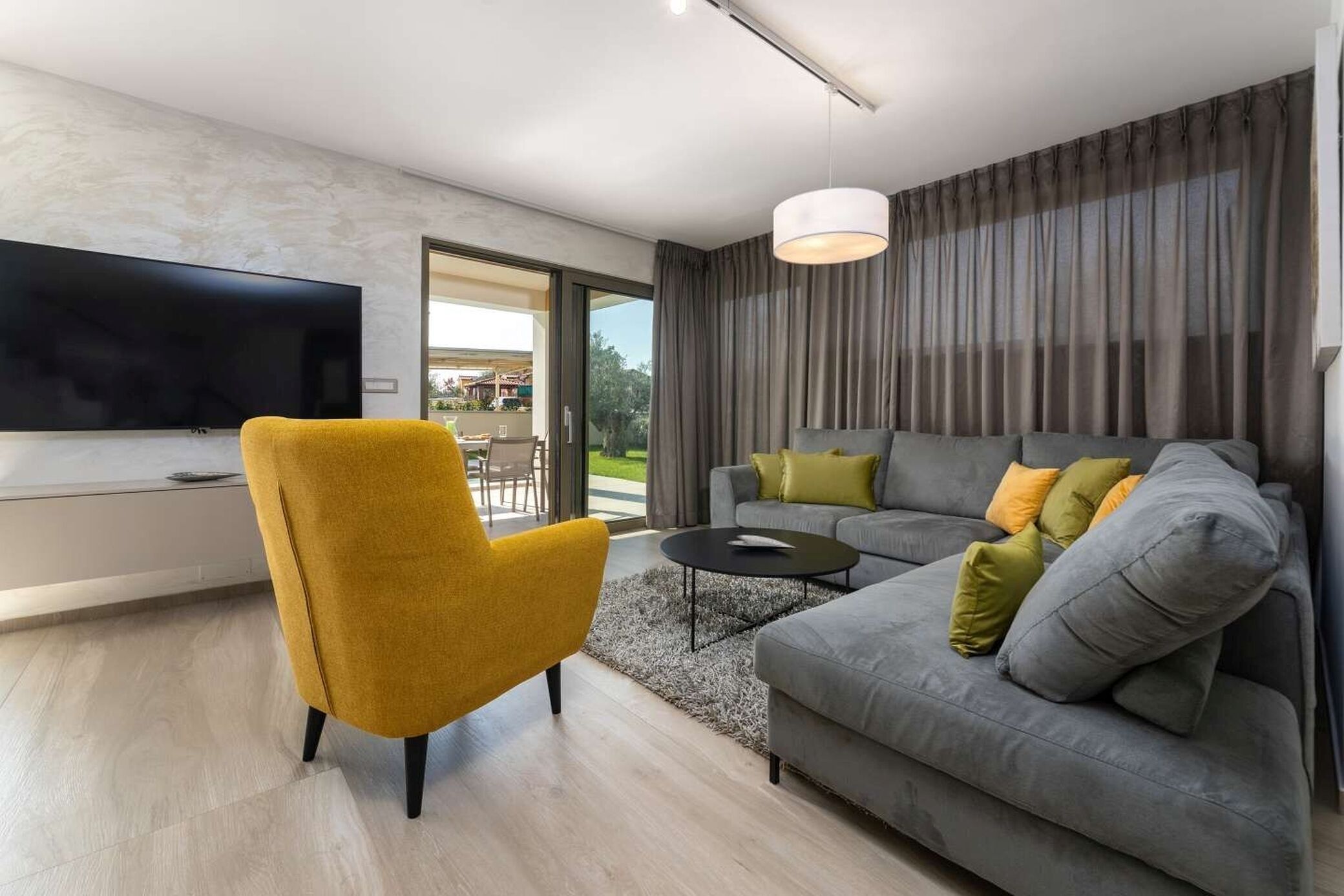 Un salon moderne doté d’un téléviseur à écran plat, d’un fauteuil jaune, d’un canapé gris avec des coussins et d’une grande fenêtre avec des rideaux.