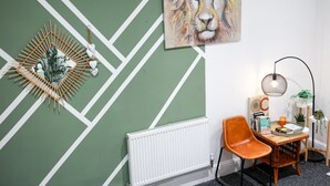 Et værelse med et grønt og hvidt geometrisk vægdesign, en hvid radiator, en træstol, et lille bord med bøger og et stearinlys, samt et hængende abstrakt billede.