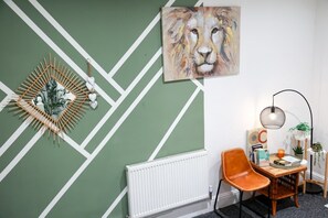 Une pièce dont le mur est décoré d’un motif géométrique vert et blanc, avec un radiateur blanc, une chaise en bois, une petite table sur laquelle sont posés des livres et une bougie, et un tableau abstrait suspendu au mur.