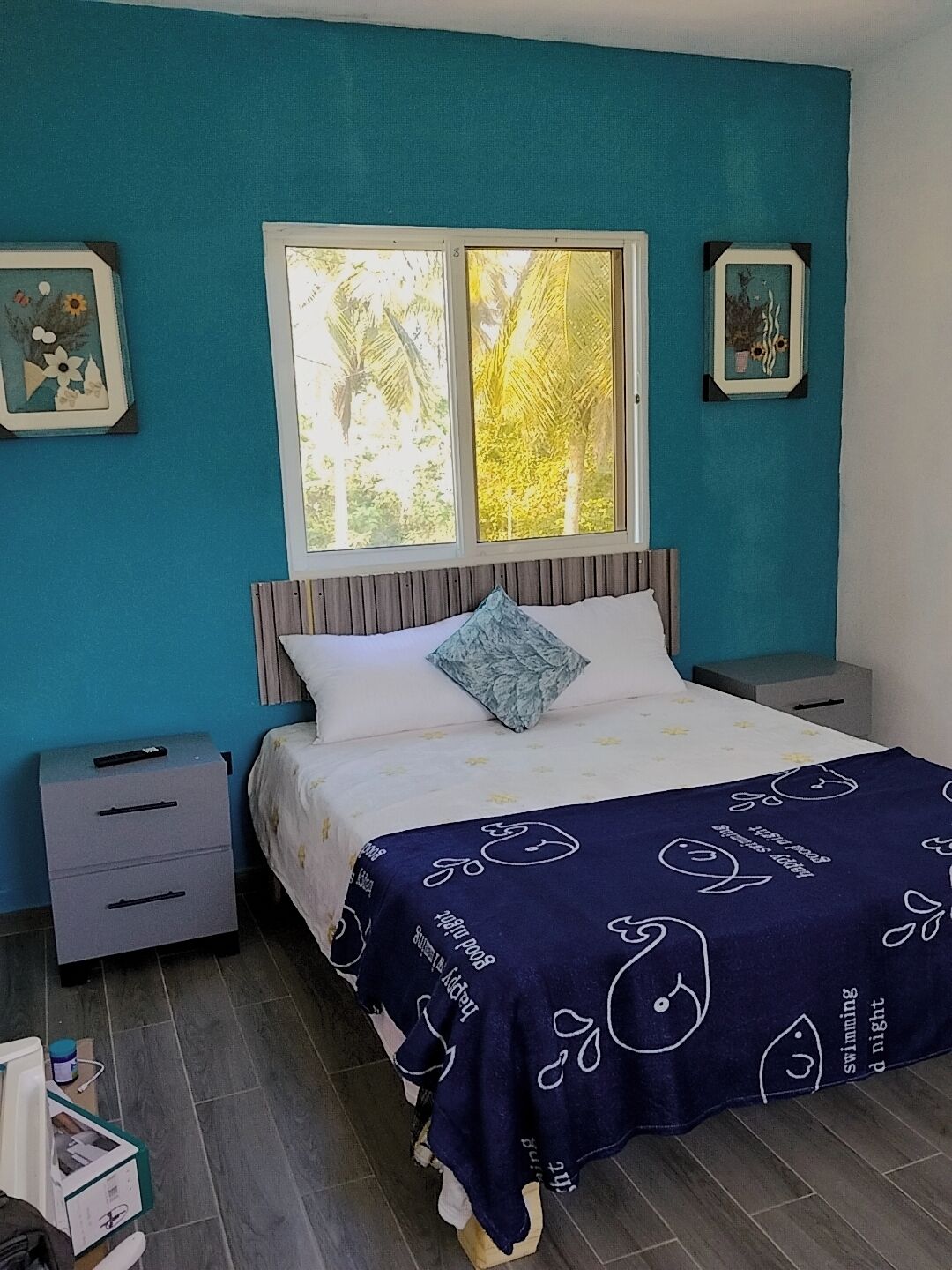 A bedroom with a bed, two bedside tables, a window with a view of trees, and framed pictures on the wall.