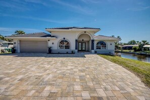 A two-story house with a covered driveway and a canal in the background. - Walk to Yacht Club Beach, Pool, Spa & Gulf Access Waterfront Escape - Cozy Corner - Roelens (Cape Coral)