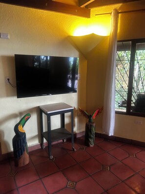 Una habitación con una televisión de pantalla plana montada en la pared, una mesita pequeña, una escultura decorativa de un pájaro y una ventana con vistas a la vegetación.