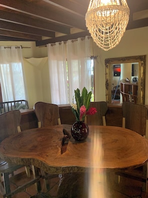 Un coin repas avec une table ronde en bois, des chaises, un vase avec des fleurs, un lustre et un grand miroir au mur.