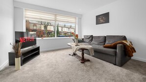 Een moderne woonkamer met een grijze bank, een salontafel, een tv waarop 'NETFLIX' staat en een raam met zonwering.